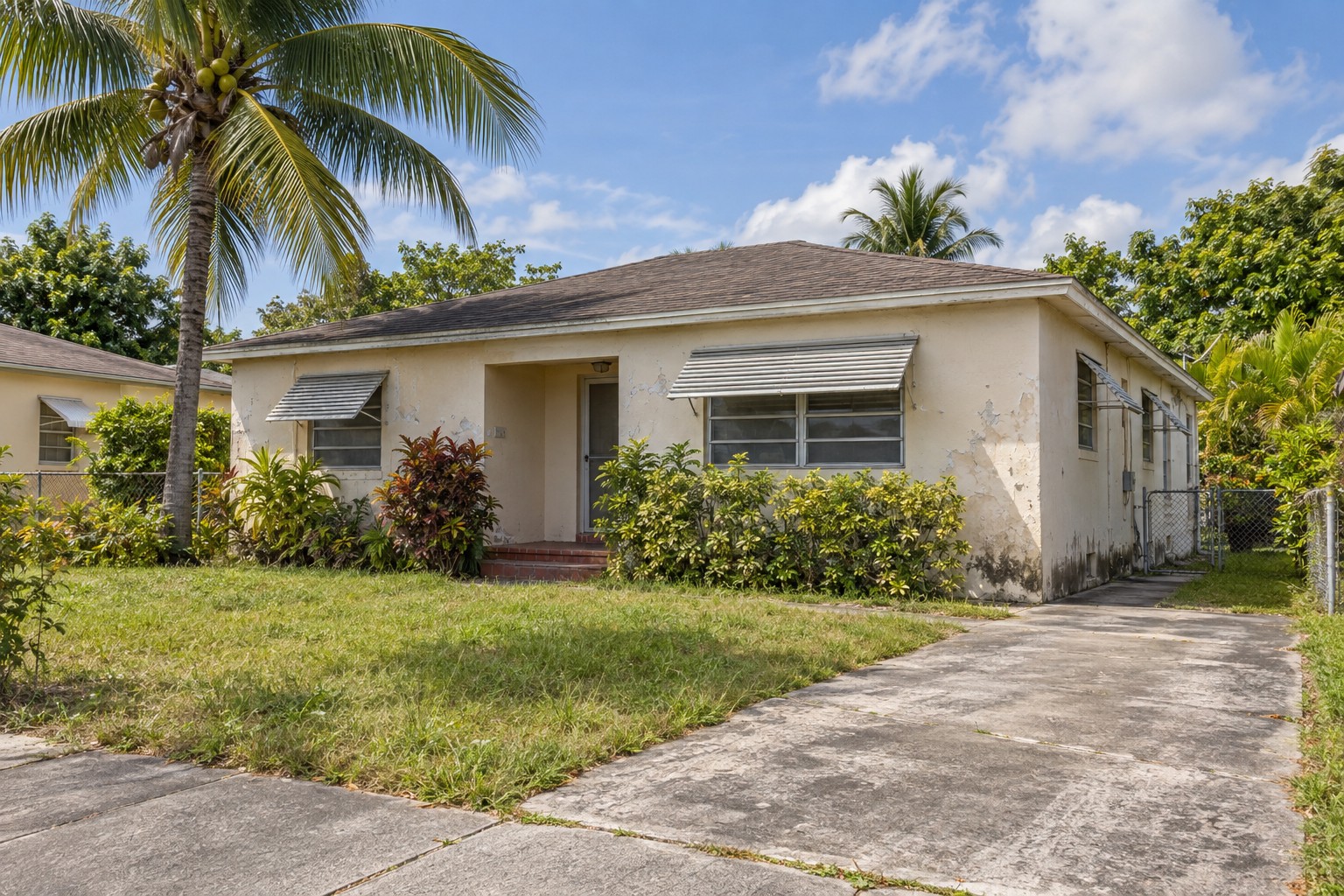 Illustrative Miami-style single-story stucco home with faded paint, aluminum window awnings, palms and a worn driveway
